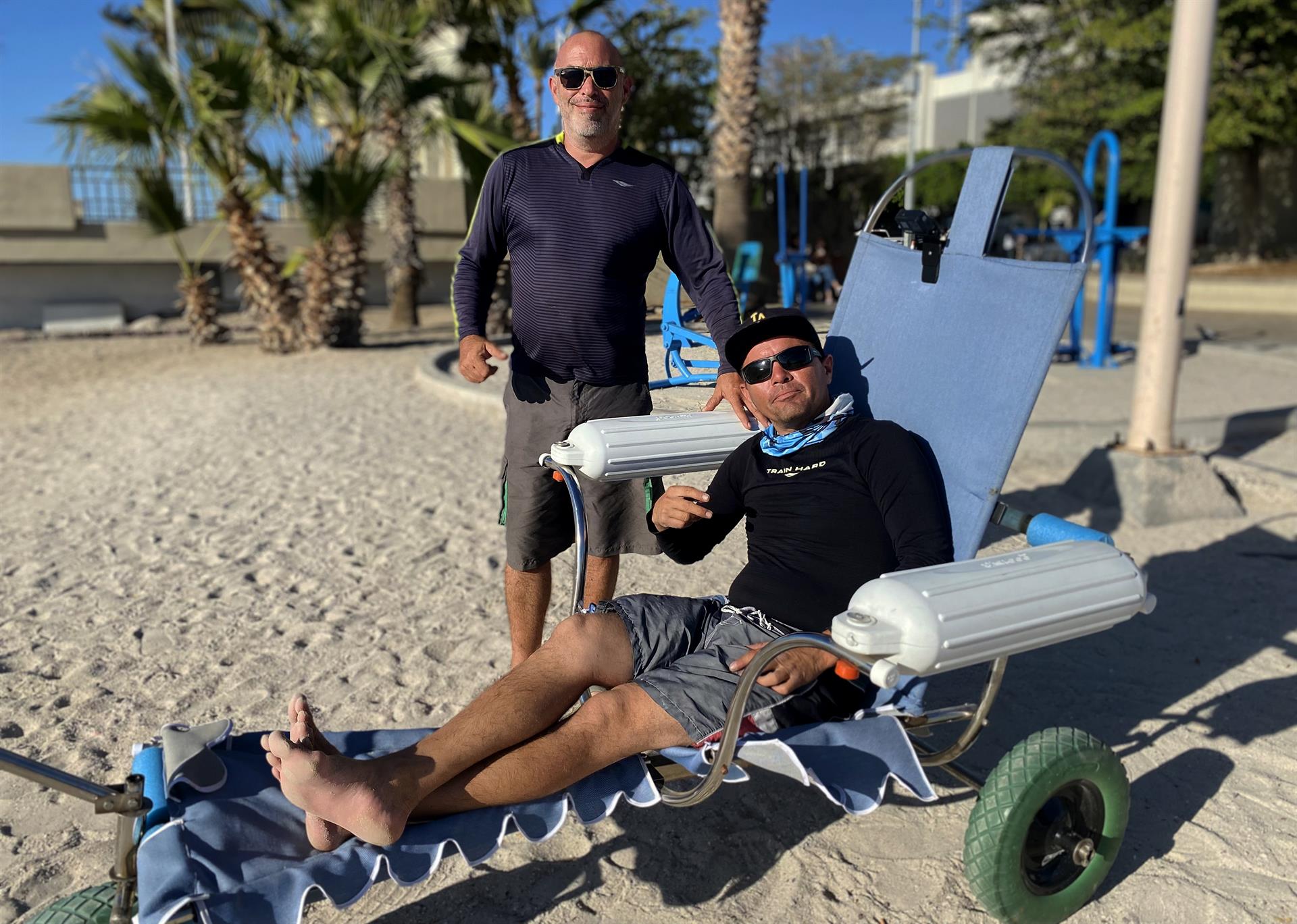 El experto en mecánica automotriz, Andrés Mata (i), y el prestador de servicios turísticos, Jairo Westle, posan junto a una silla de ruedas anfibia el 23 de abril de 2022, en una playa del balneario de La Paz (México). EFE/ Ricardo Mahatma
